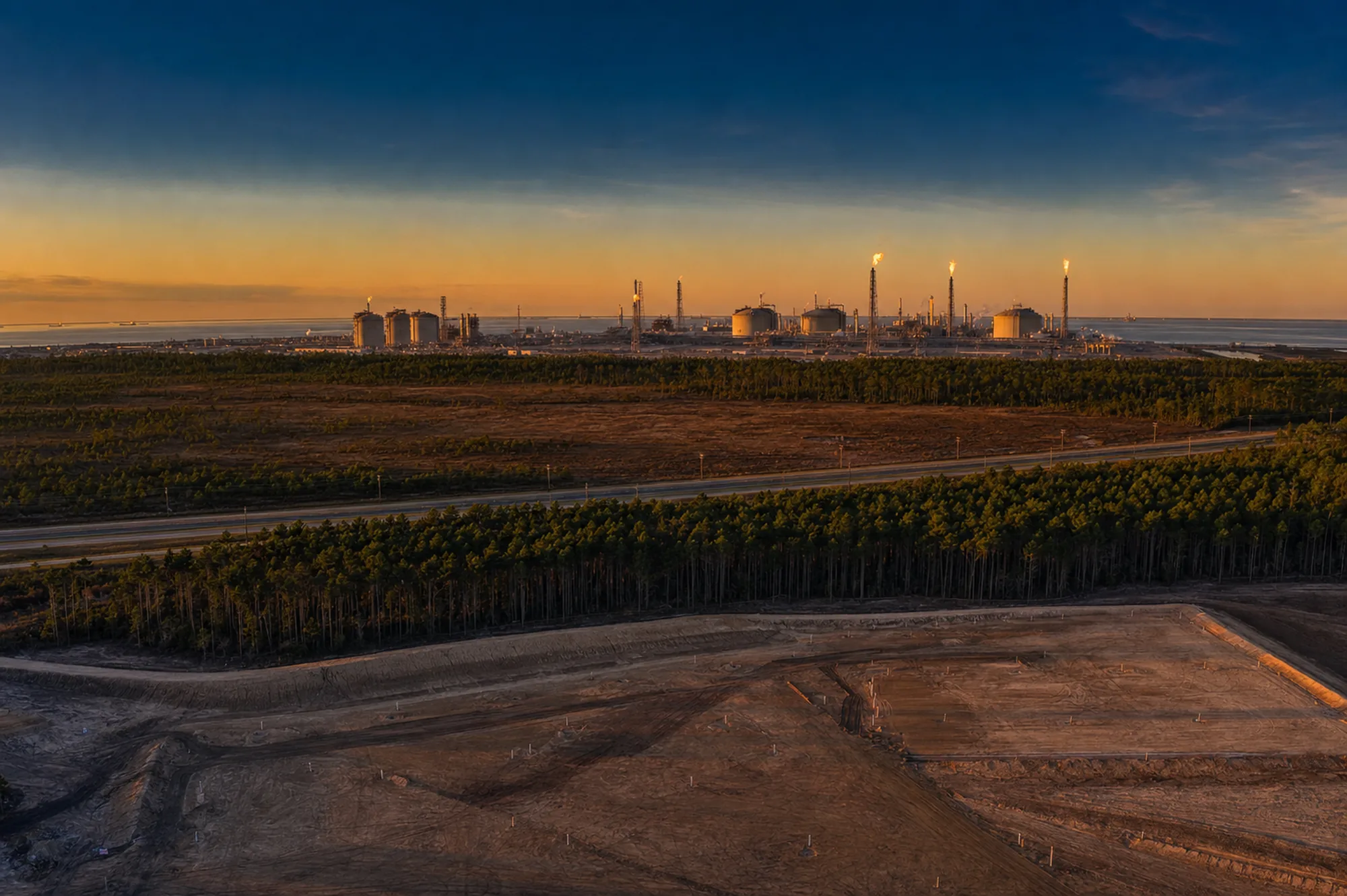 Aerial view of a graded development pad on the Texas Gulf Coast with an LNG plant on the horizon at dusk